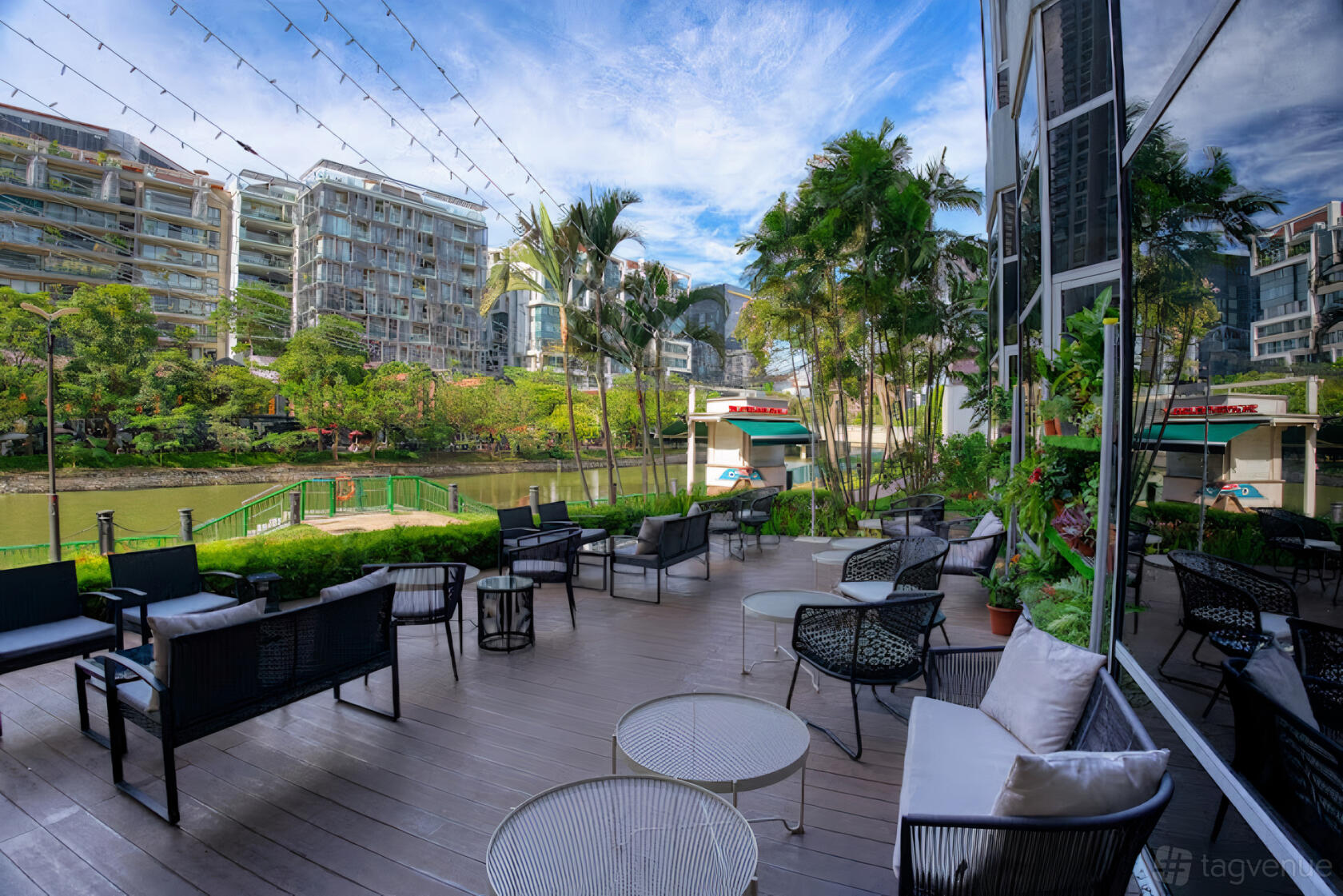 A terrace bar with outdoor seating, potted palms, and views of a river lined with buildings at The Best Brew.