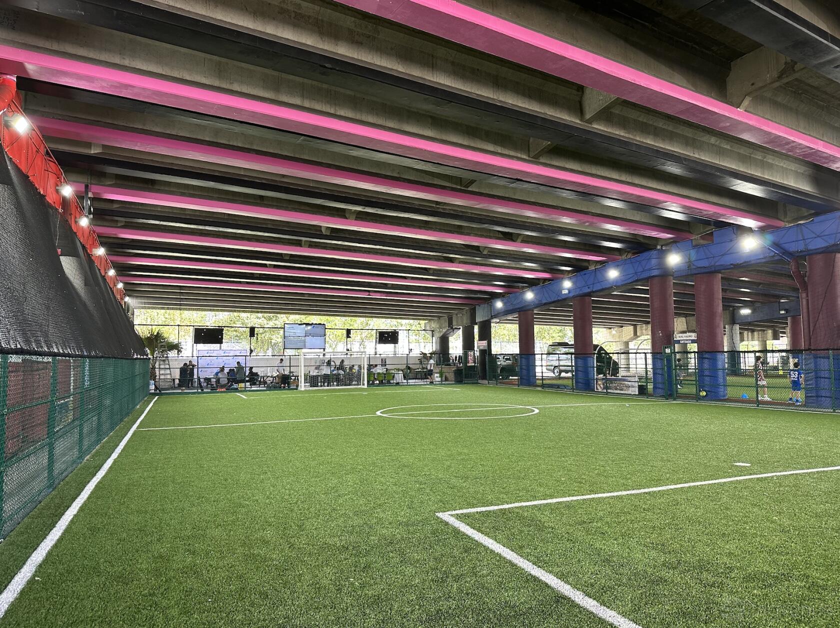 An indoor stadium with artificial turf, high ceilings with pink beams, and fenced sidelines at STADIO SOCCER.