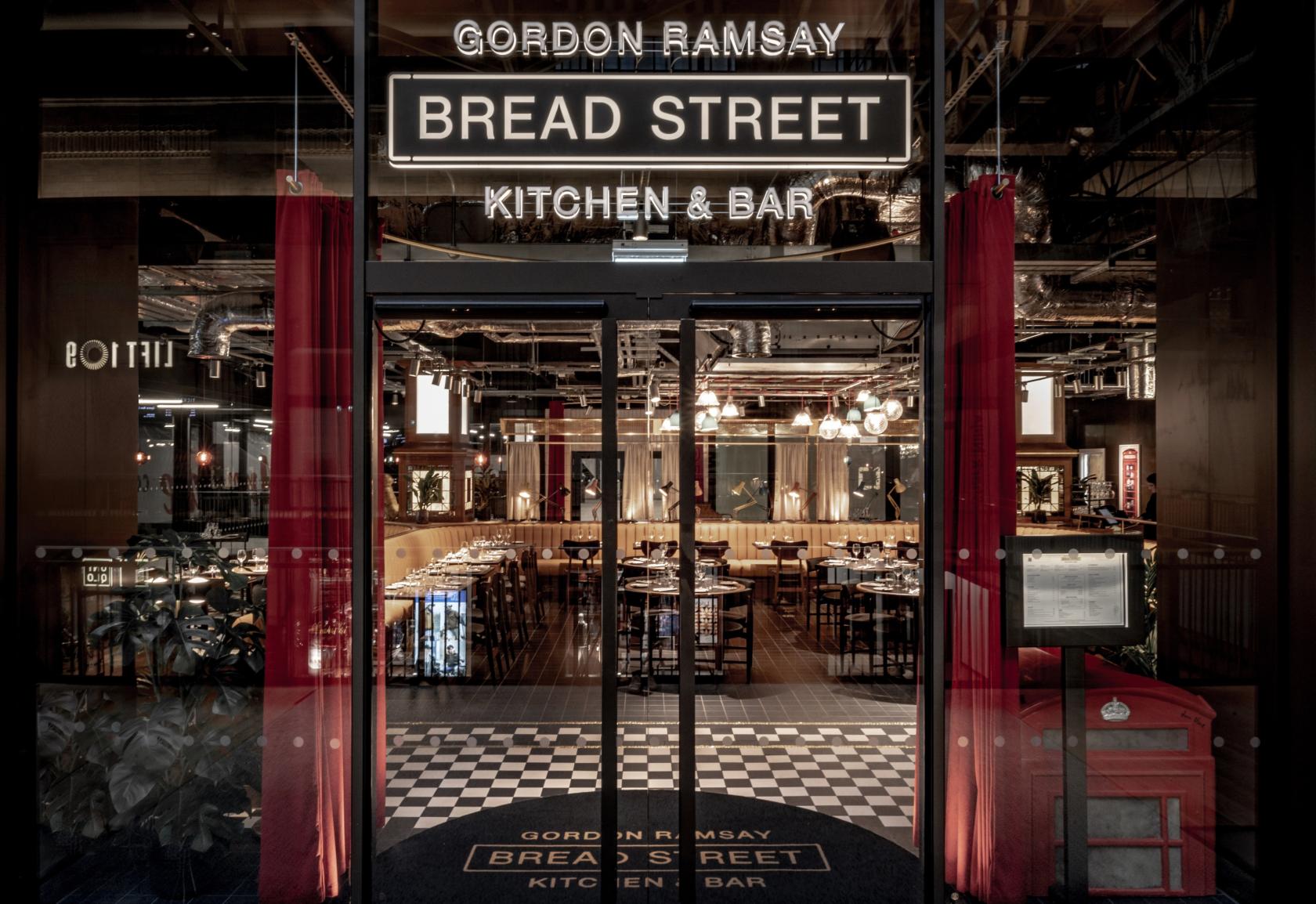 A restaurant entrance with glass doors, neon signage, and a view of wooden tables inside at Bread Street Kitchen, Battersea.