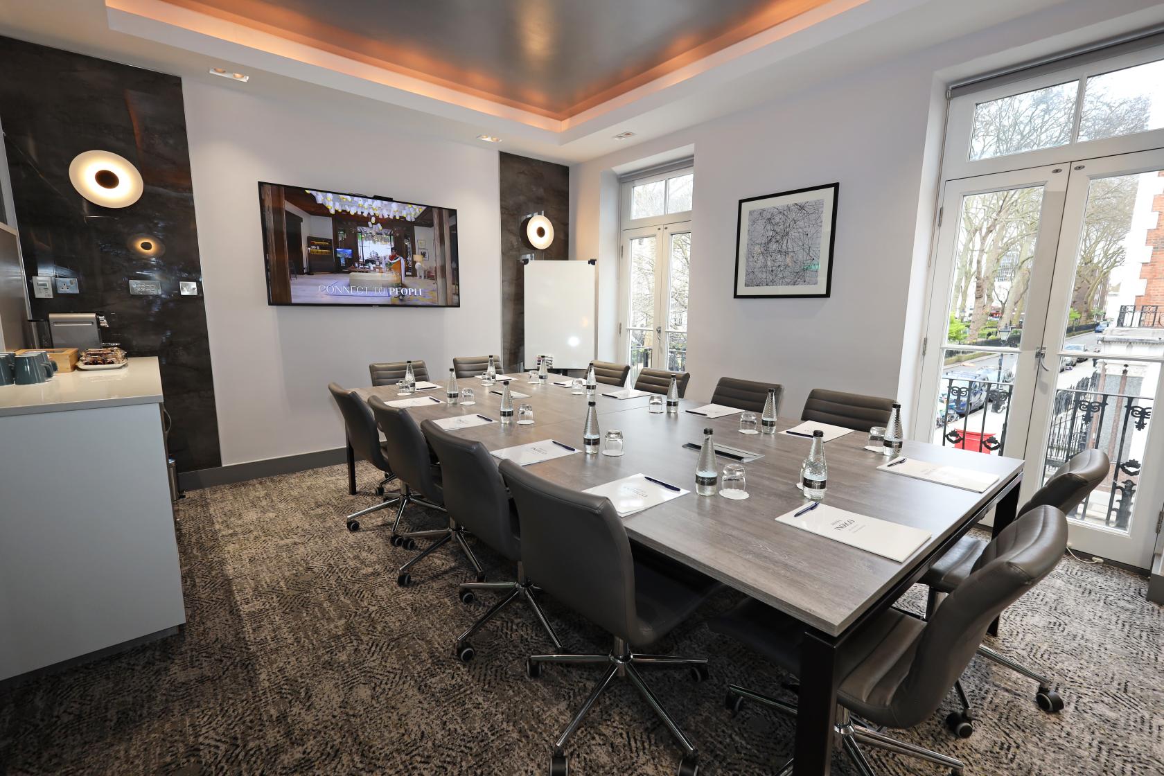 An event space with a long boardroom table, executive chairs, large windows, and wall-mounted screen at Hotel Indigo London Paddington.