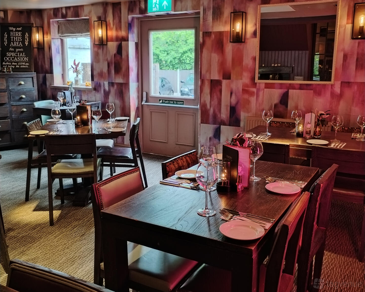 An event space in a bar with patterned wallpaper, wooden tables set with glassware, and soft wall lighting at The King William.