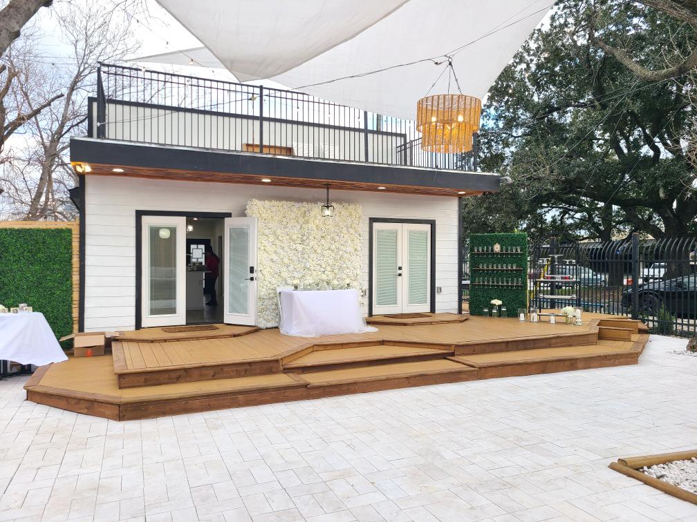 A bungalow house with a wooden deck, white double doors, and a floral backdrop at Verandas Events.