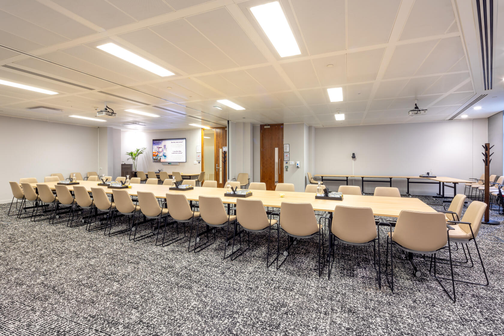 A meeting room with U-shaped tables, beige chairs, and a wall-mounted screen at Landmark: 99 Bishopsgate, London.