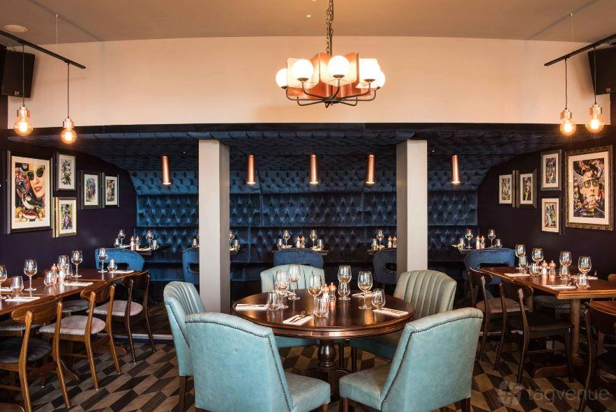 A restaurant with tufted blue banquette seating, round wooden tables, and framed artwork at Rocket.