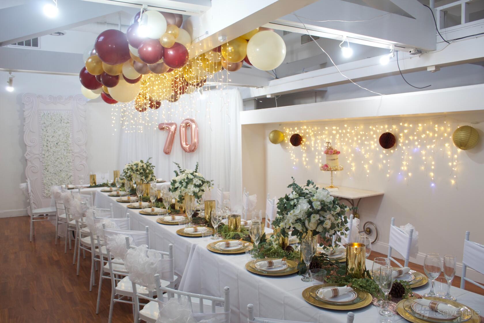 A creative event space with long banquet tables, white chairs, hanging string lights, and gold balloons at Simply Creative 2.