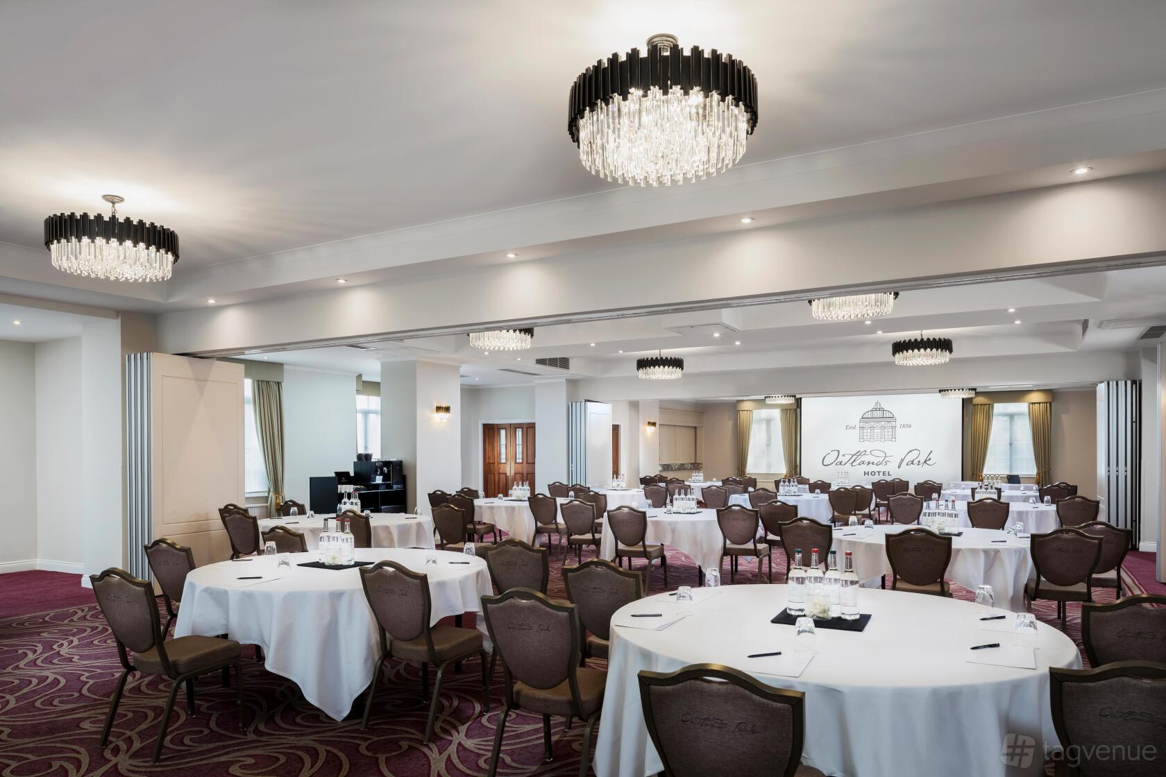 An event space with round tables, upholstered chairs, chandeliers, and carpeted floors at Oatlands Park Hotel.
