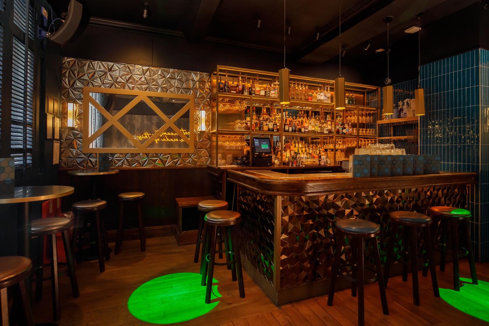 A function room with a gold-tiled bar, wooden stools, patterned wall panels, and illuminated shelves at Be At One Manchester.