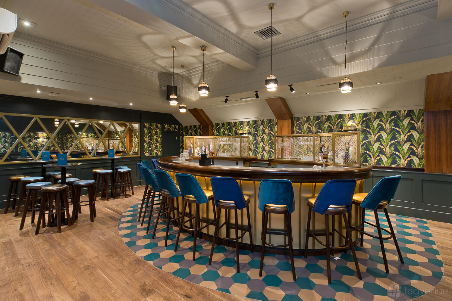 A function room with a central oval bar, blue velvet barstools, patterned tile floor, and botanical wallpaper at Be At One Manchester.