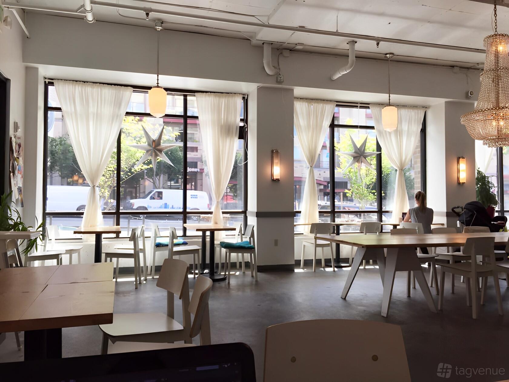A cafe with large windows, sheer white curtains, wooden tables, and hanging chandeliers at Salt and Sugar Cafe and Bakery.