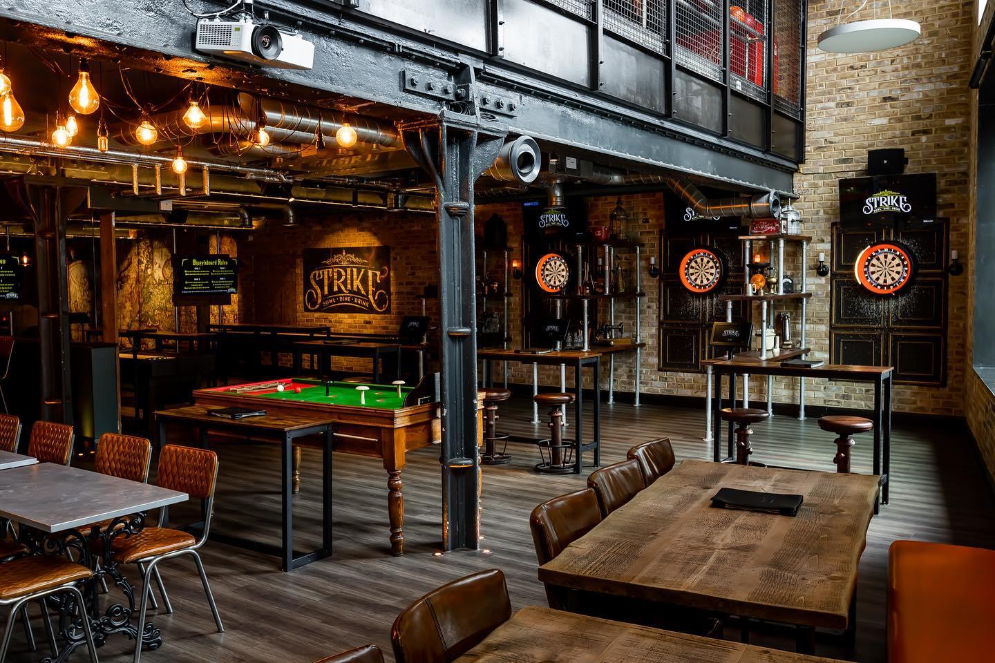 A bowling alley with exposed brick walls, industrial beams, dartboards, and communal wooden tables at Strike.