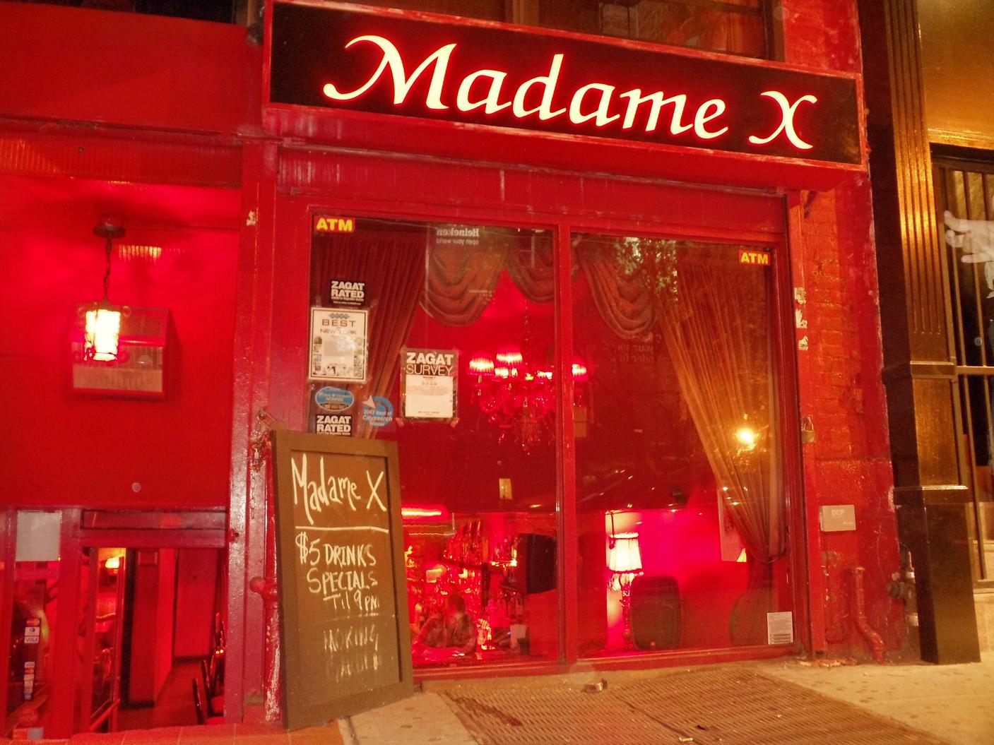 A nightclub entrance with red neon signage, red lighting, and a chalkboard drink specials sign at Madame X.