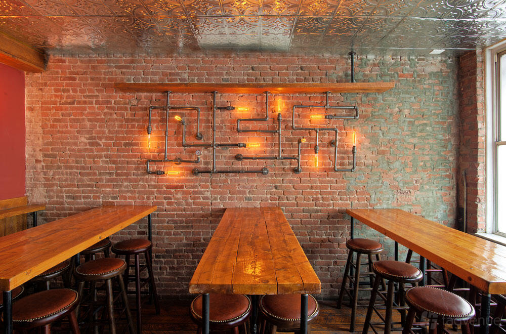 A cocktail bar with exposed brick walls, industrial pipe lighting, and wooden tables with stools at Monk McGinn's.