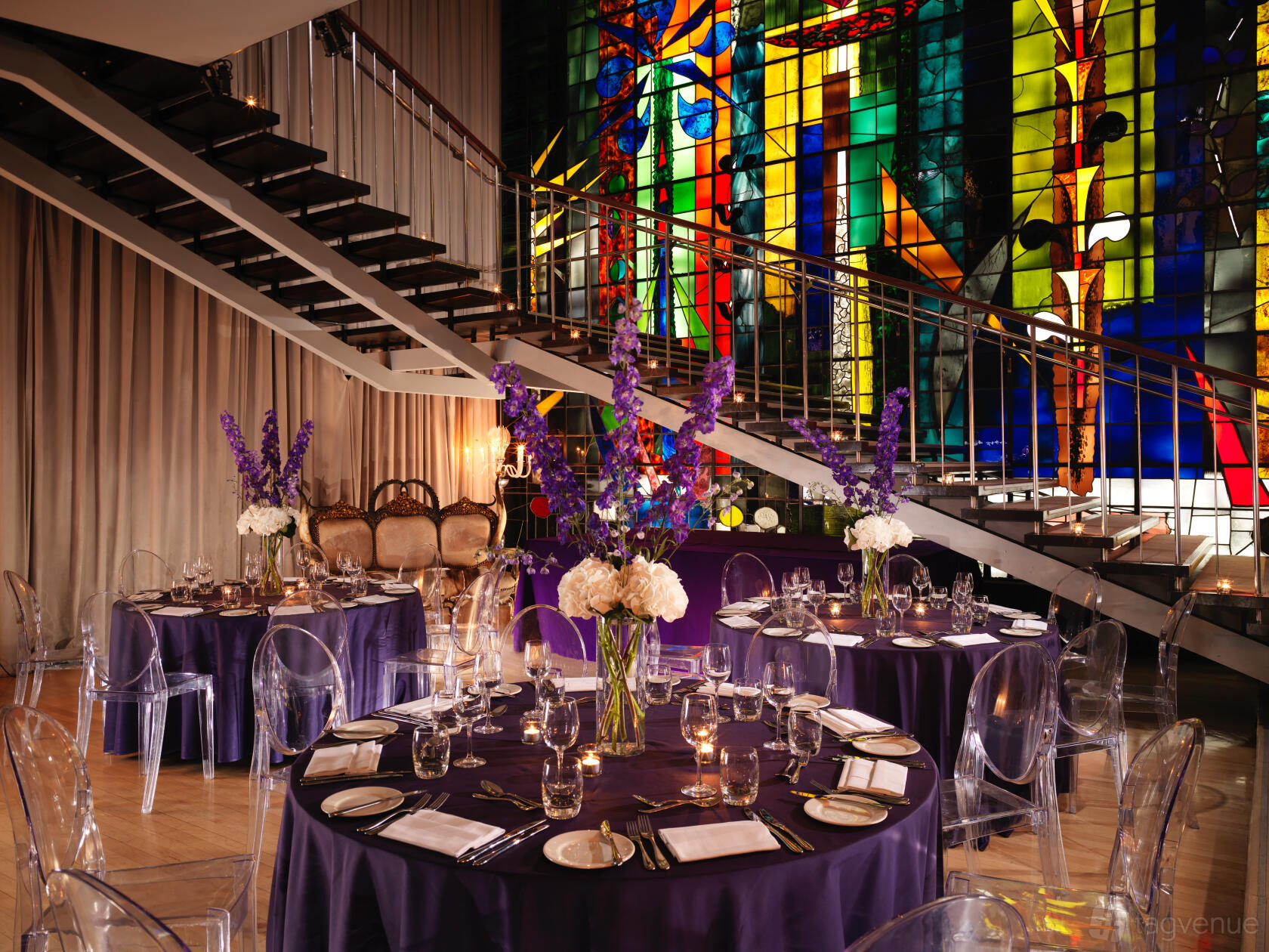 A hotel dining room with round tables draped in purple linens, tall floral centerpieces, and a stained glass backdrop at Sanderson London.