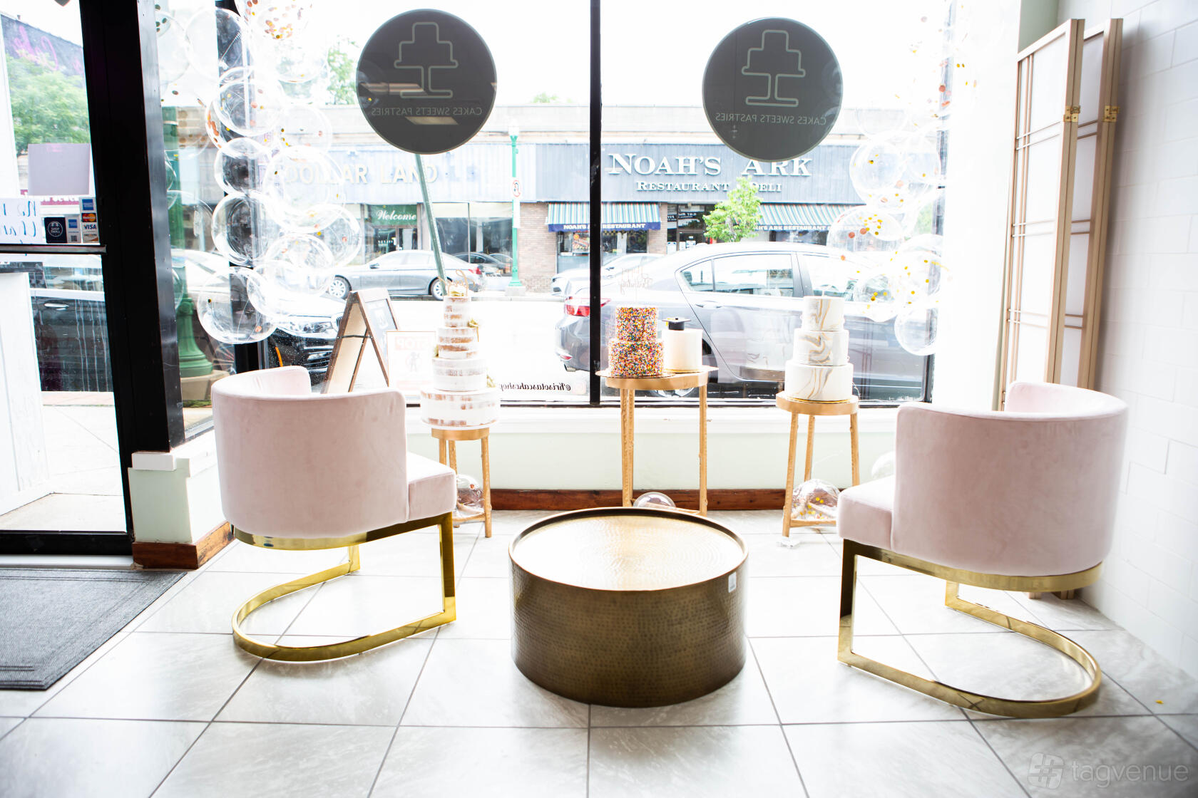 A cafe with round gold tables, blush velvet chairs, floor-to-ceiling windows, and decorative cakes at Krisota's Cake Shop.