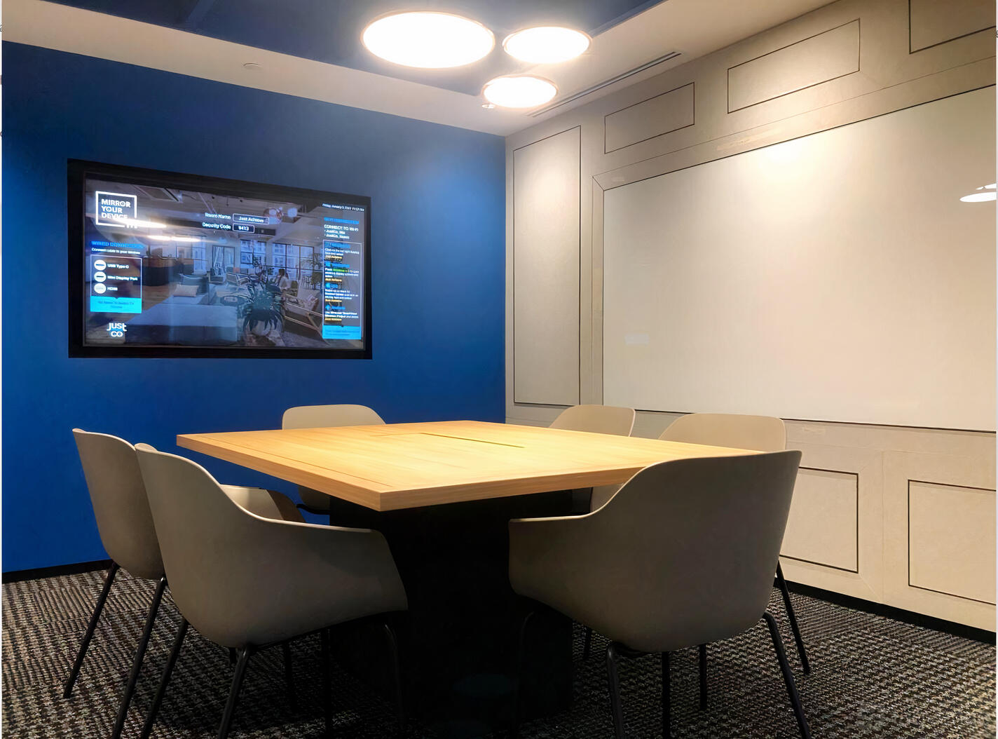 A meeting room with a wooden conference table, six white chairs, blue accent wall, and wall-mounted screen at JustCo at Collins St.