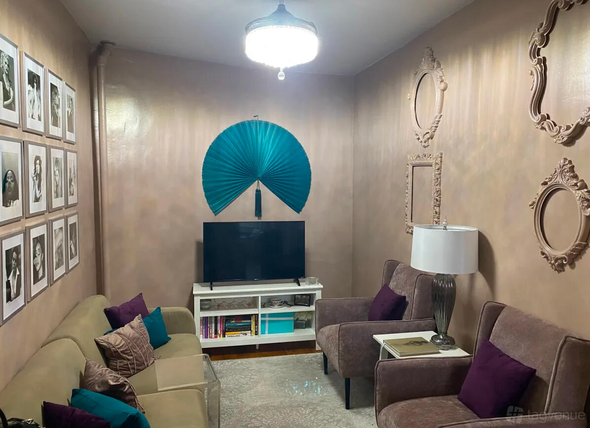 An apartment living room with a TV, gallery wall of black-and-white portraits, velvet armchairs, and decorative wall fans at Quirky Brooklyn Apartment.