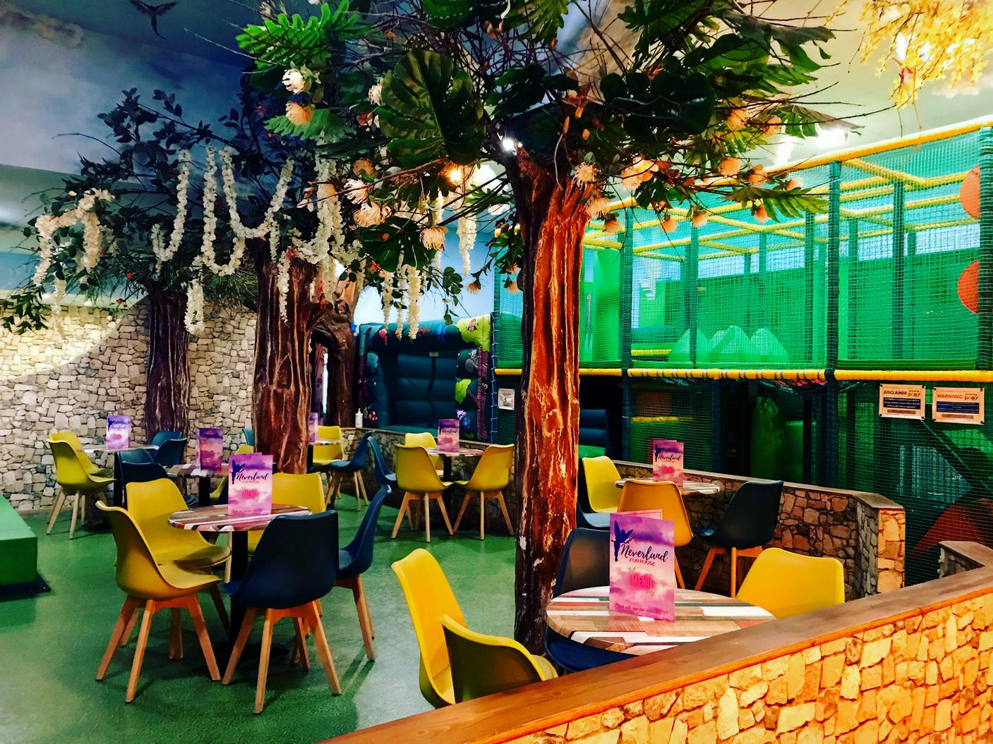 An event space with artificial trees, stone-patterned walls, colorful chairs, and a large indoor play structure at Neverland Fun House.