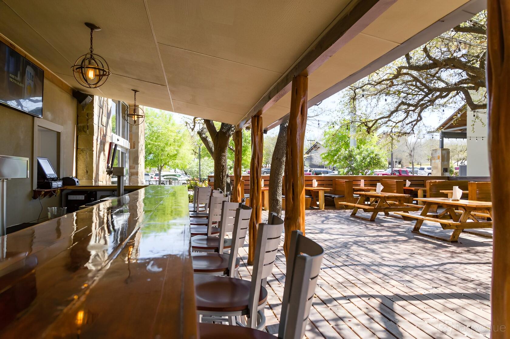 An outdoor restaurant patio with a polished bar counter, wooden chairs, and picnic tables under leafy trees at Hops and Thyme.