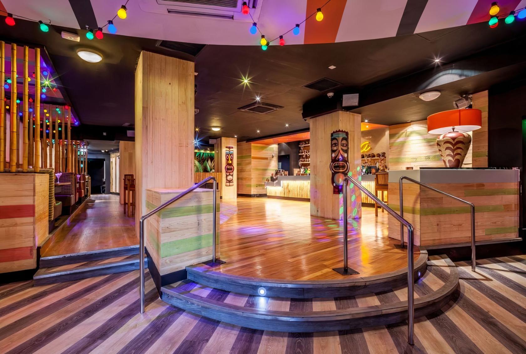 A nightclub bar with colorful string lights, wooden pillars, bamboo accents, and a tiki statue at PRYZM Brighton.