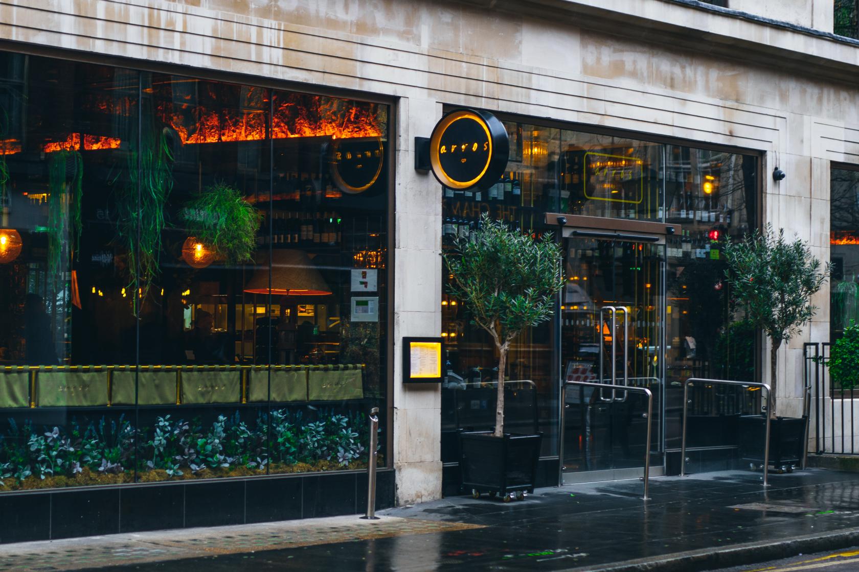 A restaurant with large street-facing glass windows, hanging greenery, and glowing orange lighting at ArrosQD.