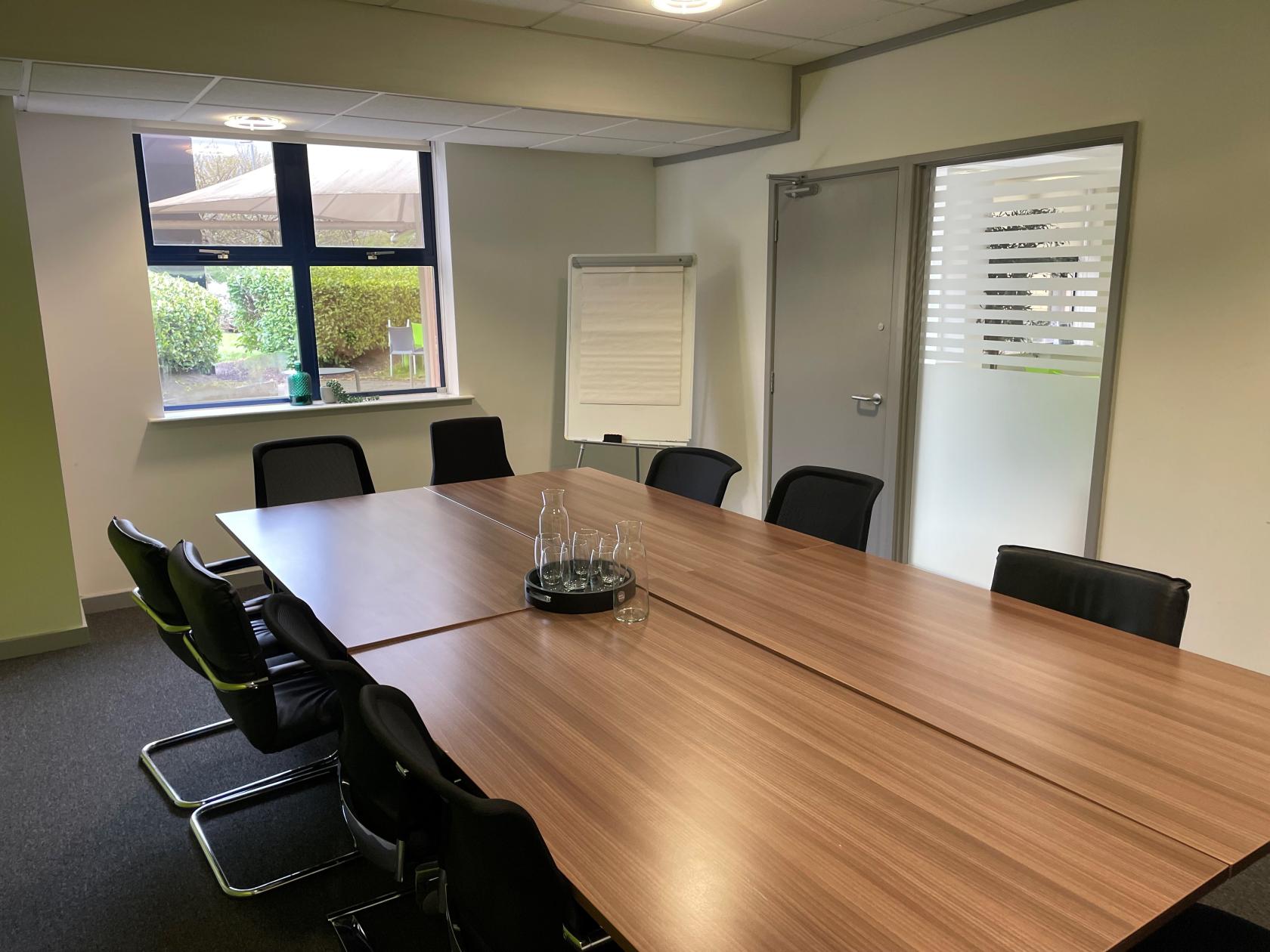 A meeting room with a large wooden table, black chairs, a flip chart, and a window at Flexico - YBN - Gateshead.