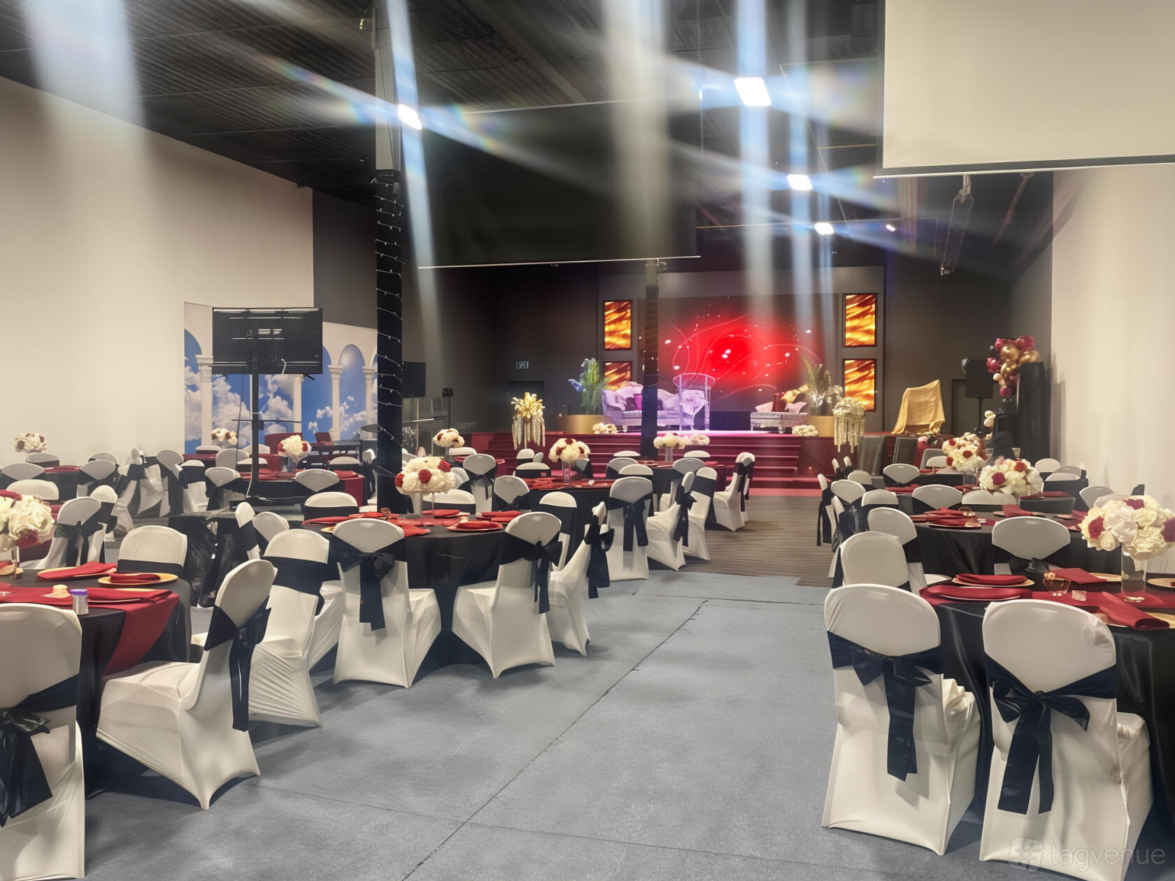 An event venue with round tables dressed in white linens, black chair sashes, and a stage with decorative lighting at Metropolitan Event Centre.