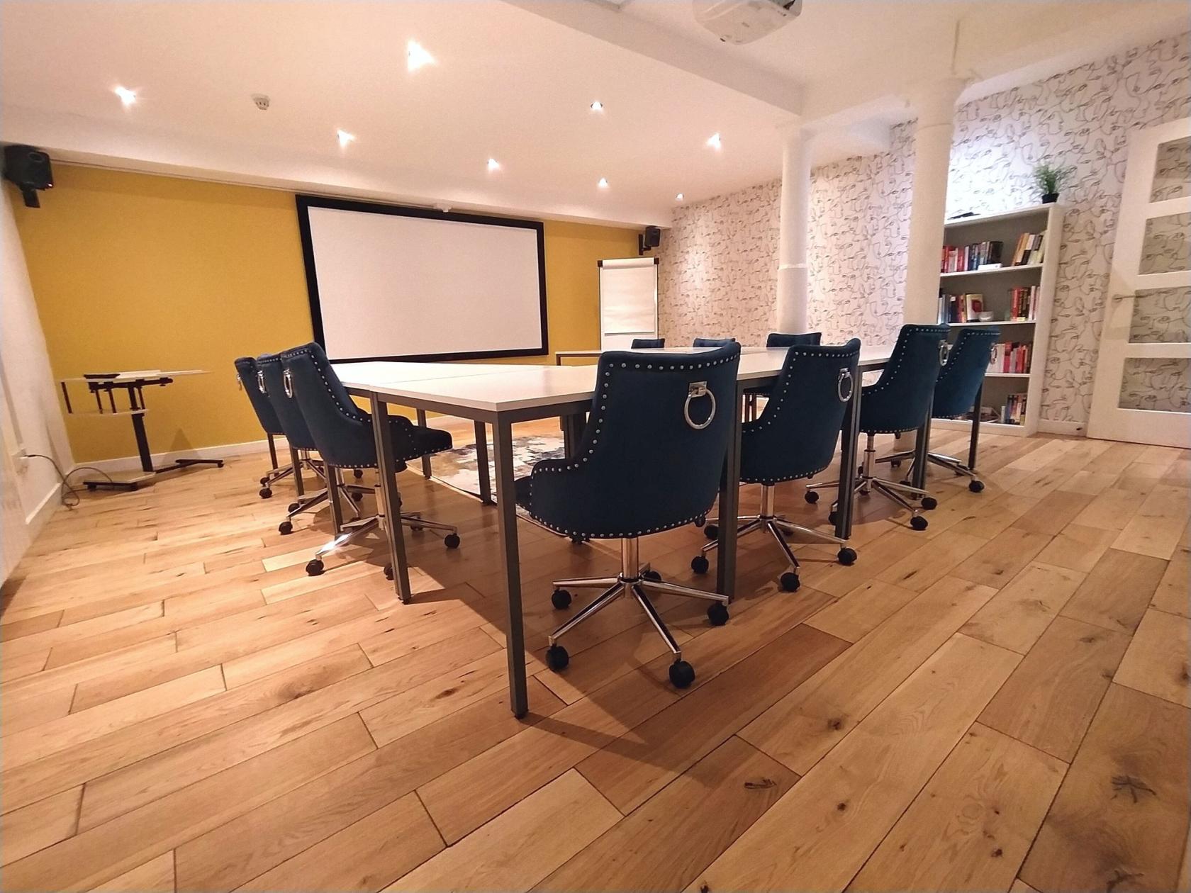 A meeting room with a large screen, navy chairs on wheels, light wood flooring, and built-in bookshelves at Collabor8te.