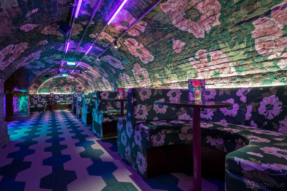 A bar with arched exposed brick ceilings, floral upholstered booths, and purple neon lighting at Blame Gloria Covent Garden.