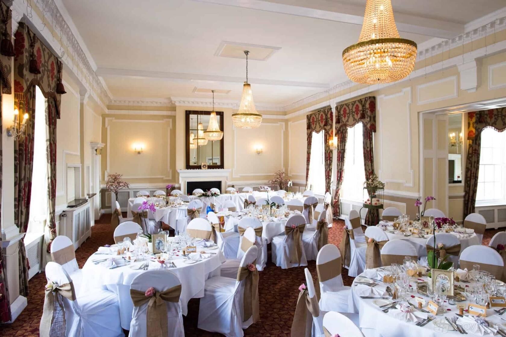 A hotel banquet hall with round tables covered in white linens, chandelier lighting, and large windows at The Maids Head Hotel.