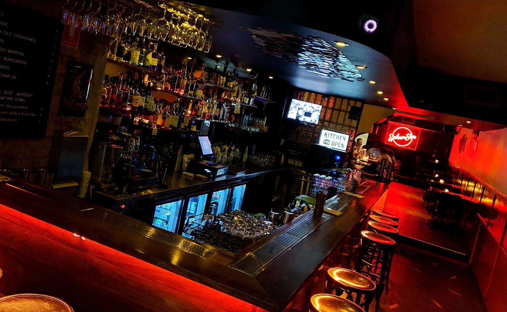 An event space in a bar with a long wooden counter, backlit shelves of liquor, and red neon lighting at Spleen Bar.