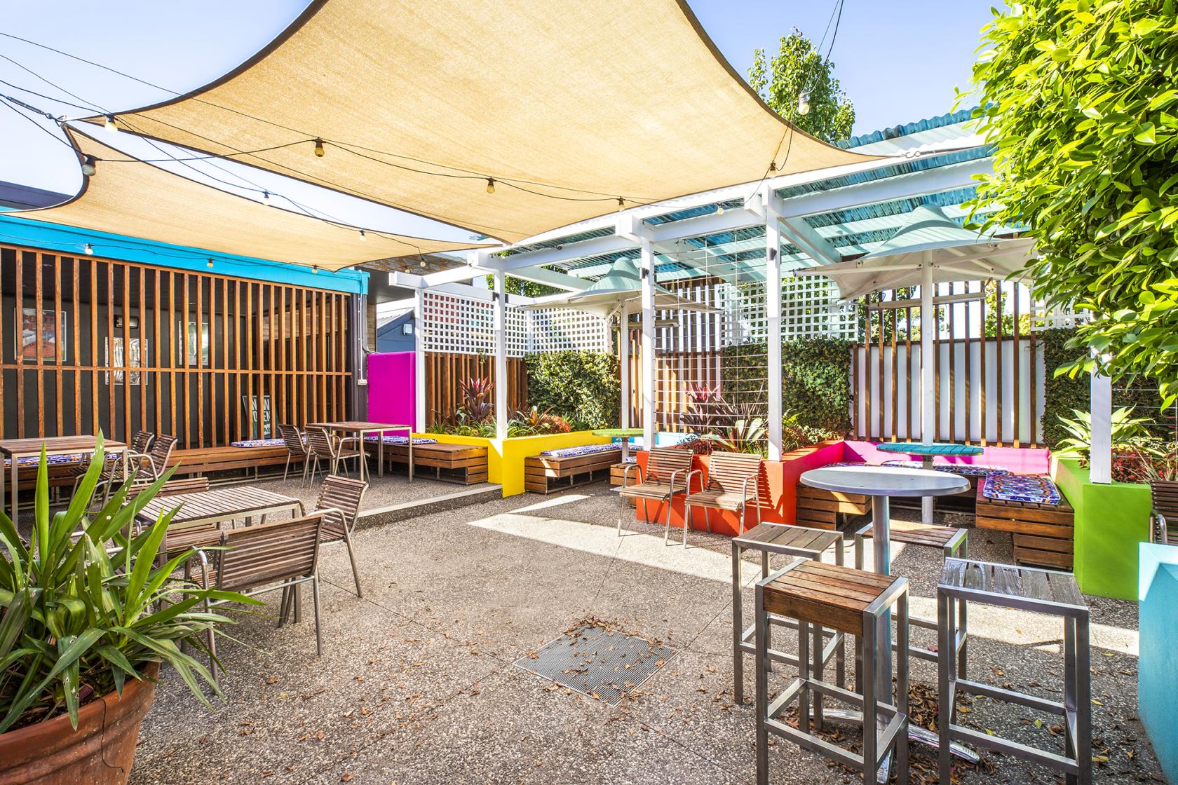 A pub beer garden with wooden tables, colorful benches, shade sails, and surrounding greenery at Keysborough Hotel.