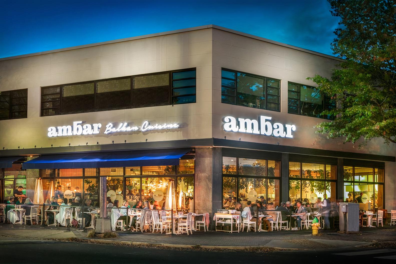 A restaurant with large windows, outdoor dining tables, and illuminated signage at Ambar Clarendon.