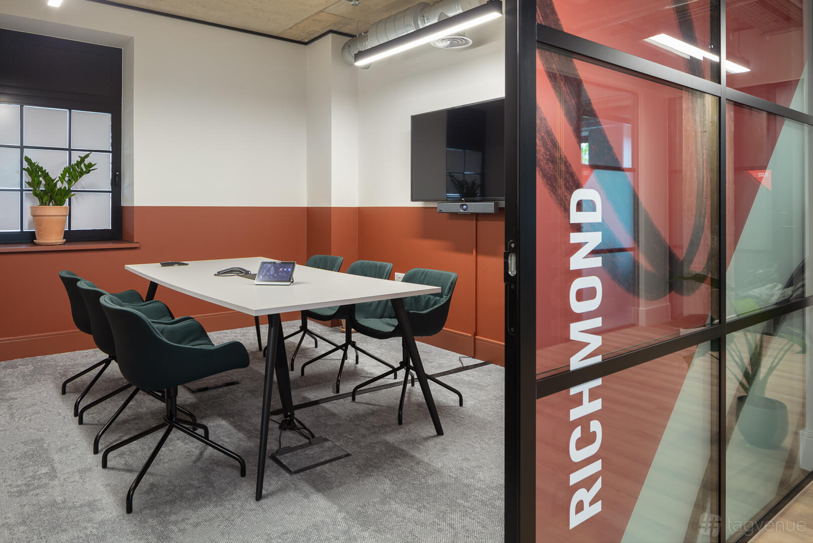 A meeting room with a rectangular table, dark green chairs, wall-mounted screen, and large window at Riley Studios by Spacemade.