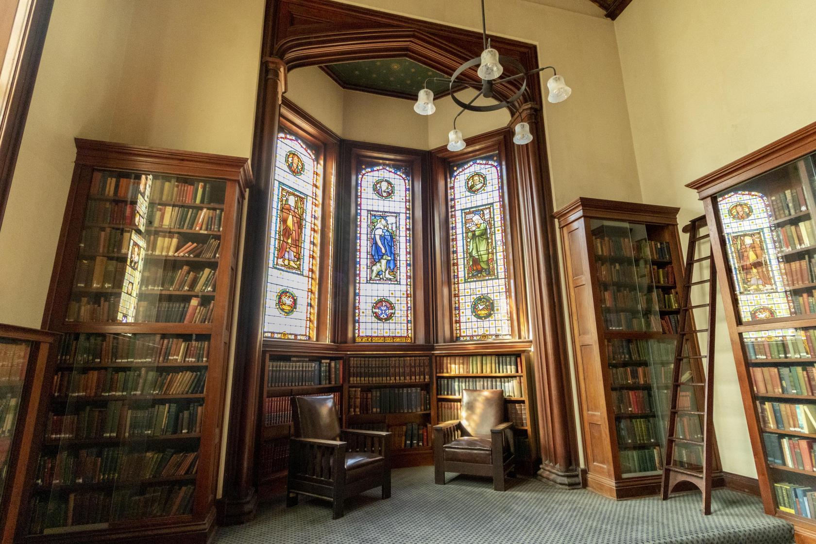A university library with stained glass windows, tall wooden bookshelves, and leather armchairs at St Andrew's College University of Sydney.