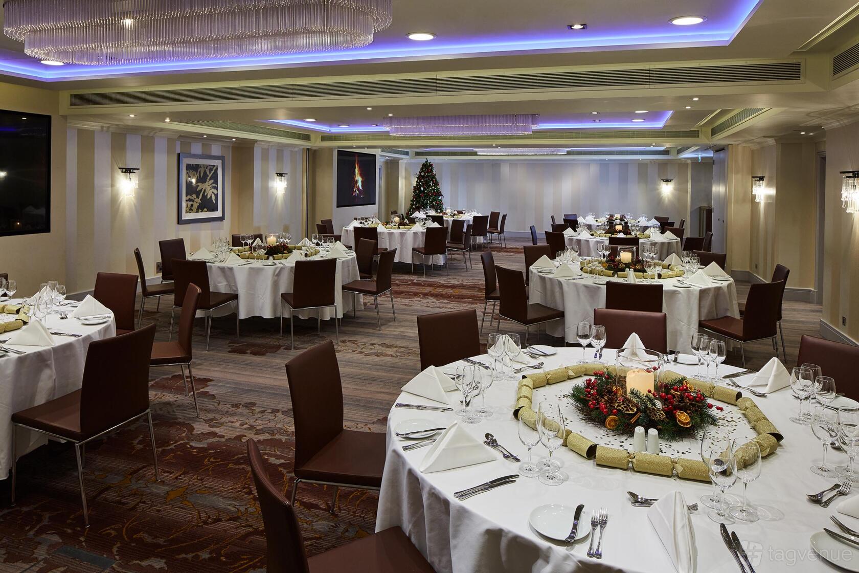 A hotel banquet room with round tables, brown chairs, white linens, and holiday decorations at Meetings & Events Venues.