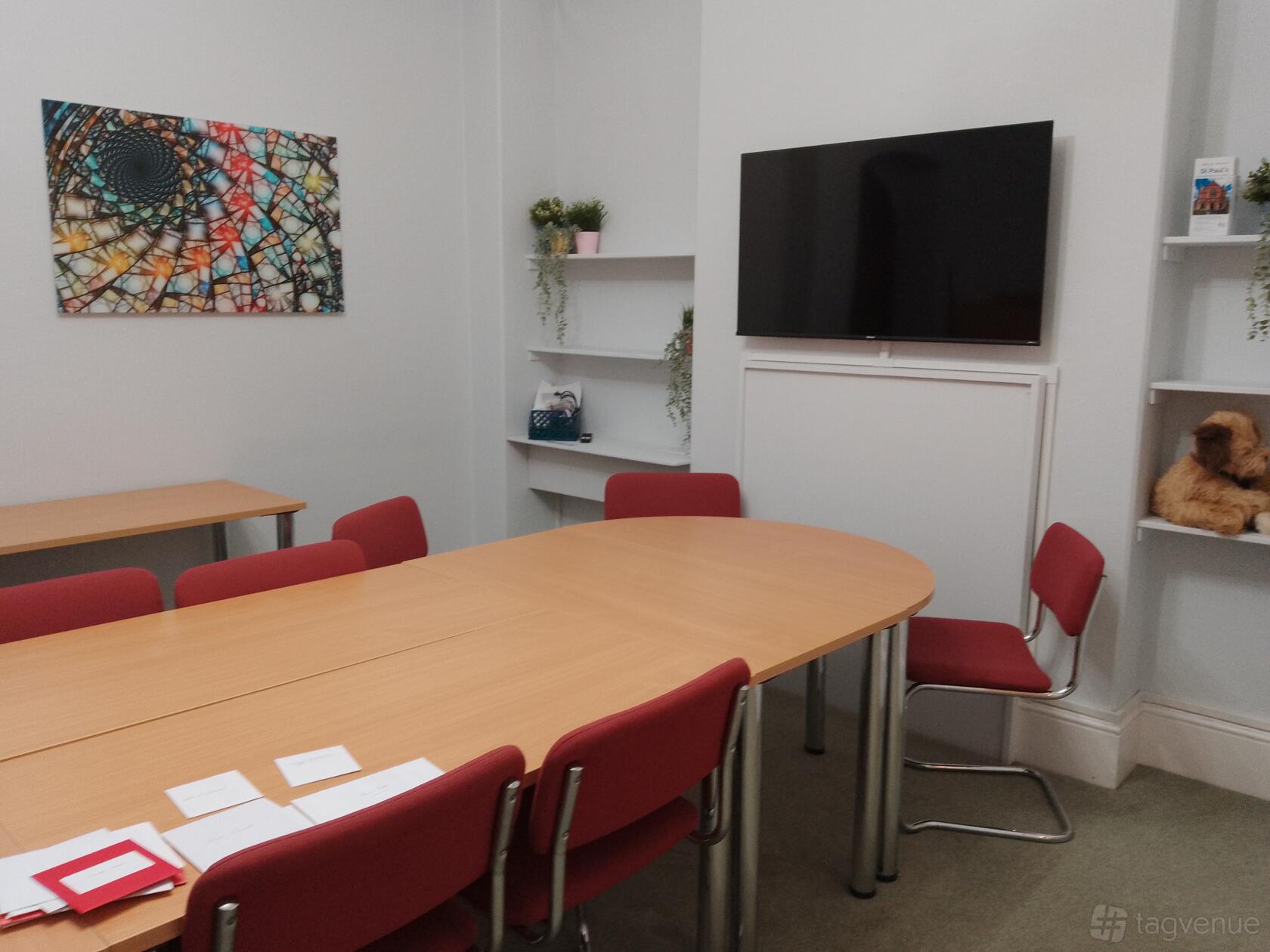 A church meeting room with a wooden oval table, red chairs, wall-mounted TV, and decorative shelves at St Paul's UR Church and halls Croydon.