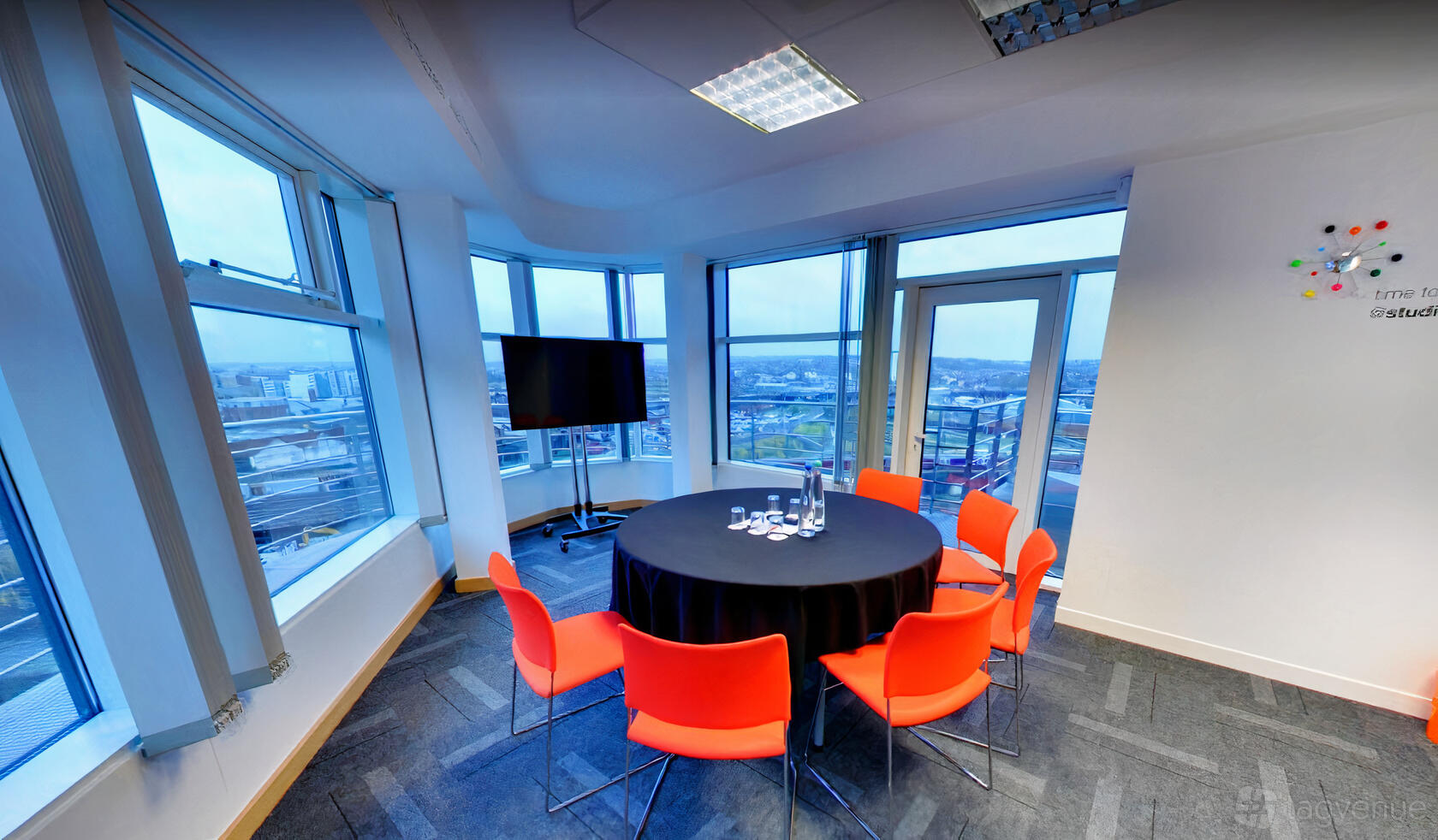 A meeting room with floor-to-ceiling windows, a round table with a black cloth, and red chairs at The Studio Leeds.