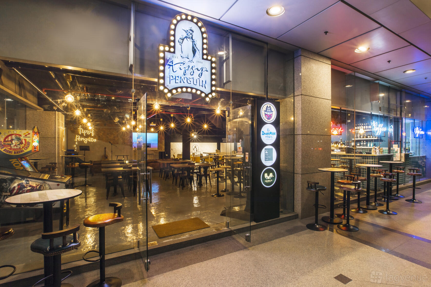 A pub with floor-to-ceiling glass frontage, high tables, and exposed bulb lighting at Tipsy Penguin.