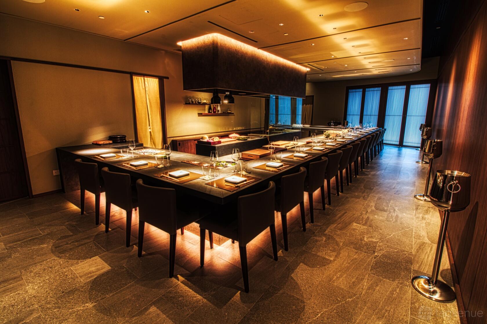 A restaurant with a long central chef’s counter, upholstered chairs, and warm recessed lighting at Kappou.