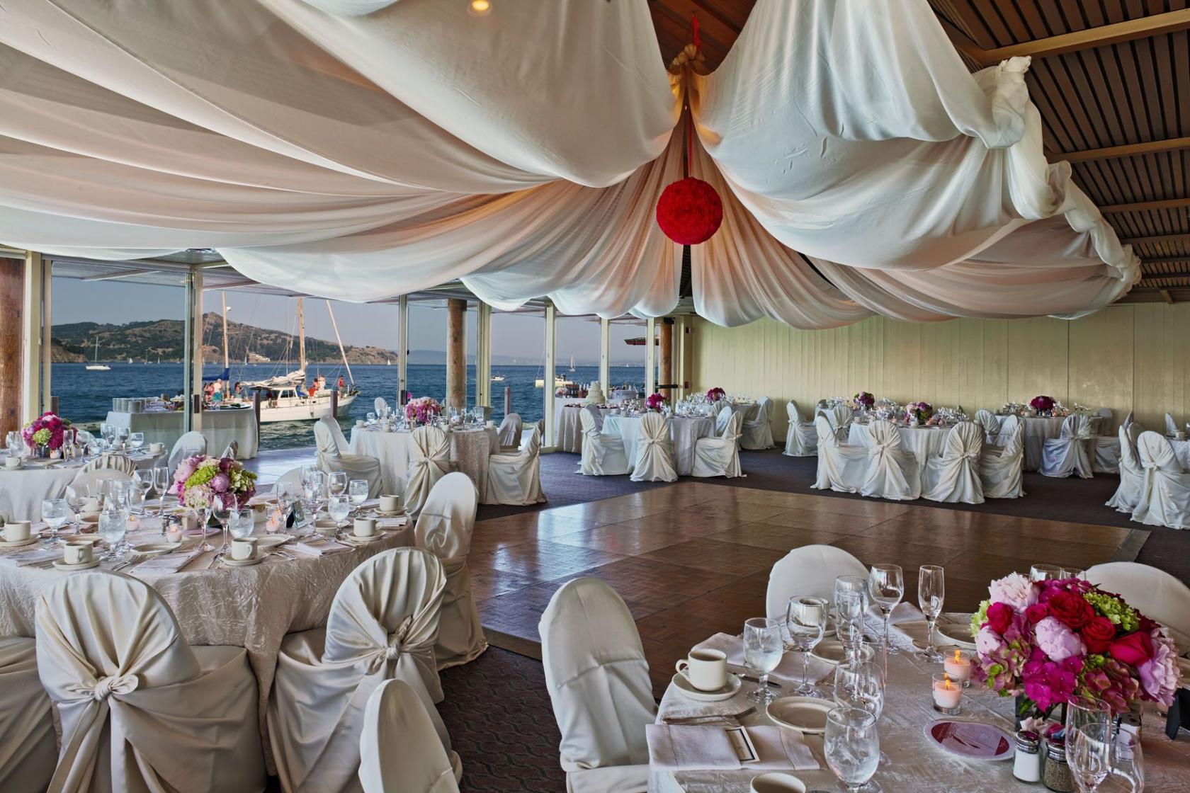 An event space with draped fabric ceiling, round tables with white linens, and waterfront views at The Spinnaker.