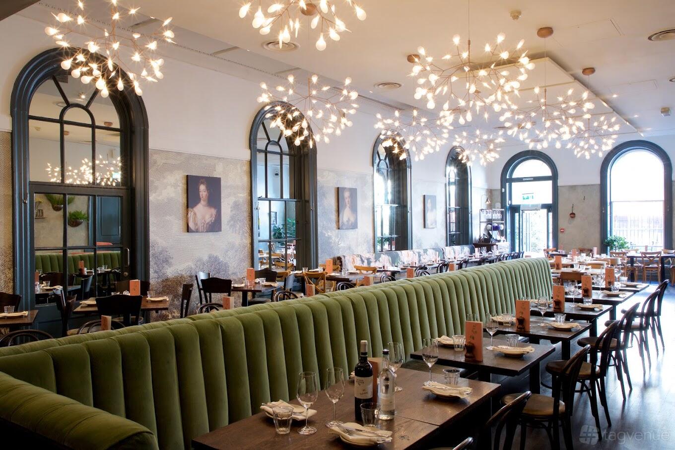 A restaurant with green velvet banquette seating, large arched windows, and modern chandeliers at Brasserie Blanc Cheltenham.