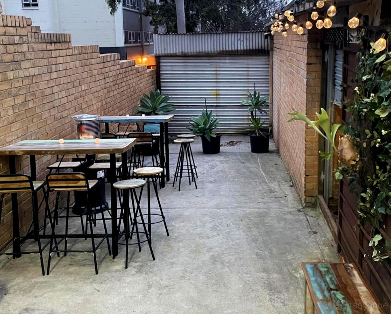 A pub terrace with high wooden tables, metal stools, potted plants, and exposed brick walls at Twang Brewing.