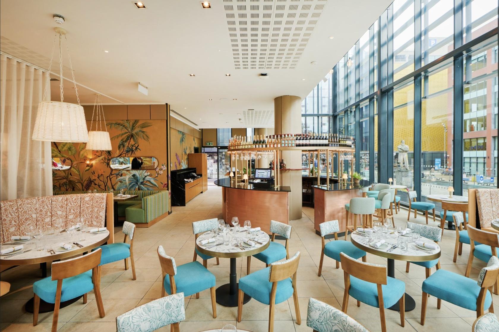 An event space in a restaurant with floor-to-ceiling windows, teal chairs, round tables, and hanging lamps at Gino D'Acampo Manchester.