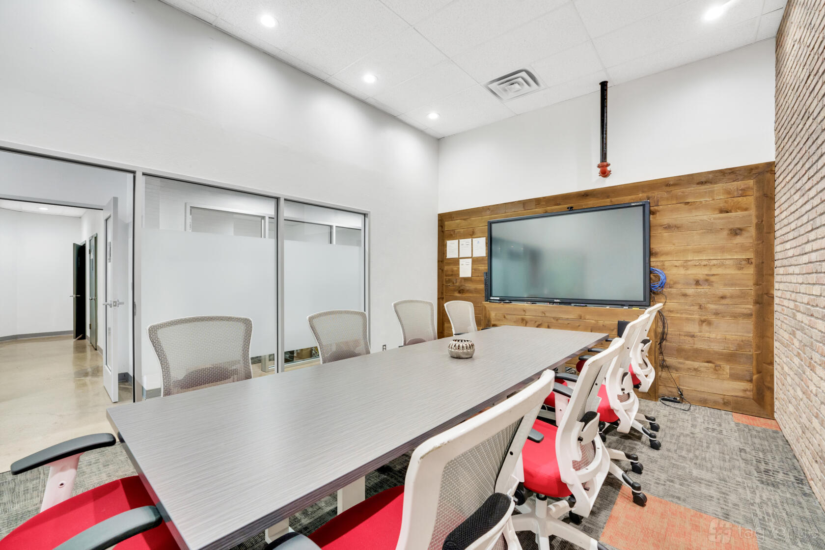 A conference room with a long table, red-accented chairs, glass walls, and a wall-mounted display at The Drawing Board.