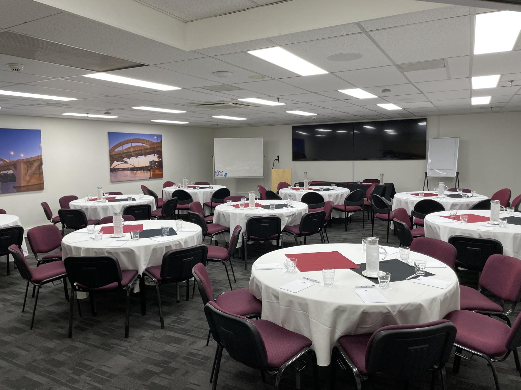 An event space with round tables covered in white linens, burgundy chairs, and a wall-mounted screen at George Williams Hotel.