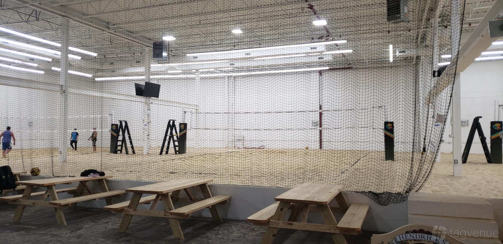A warehouse with three sand volleyball courts, bright overhead lights, and wooden picnic tables at Giant warehouse with 3 sand volleyball courts.