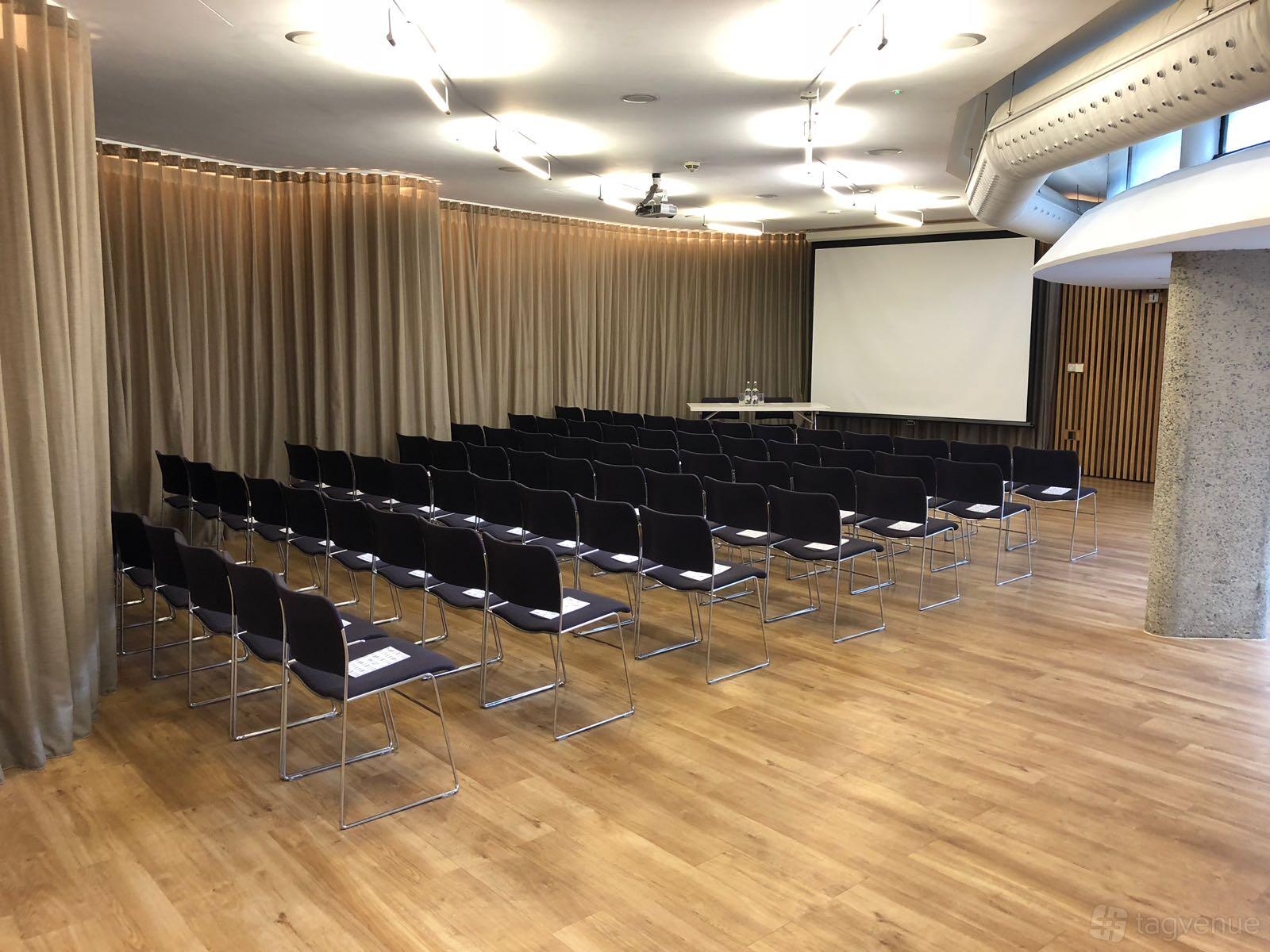 An event space with rows of black chairs, wood flooring, ceiling lights, and floor-to-ceiling curtains at London Museum Spaces.