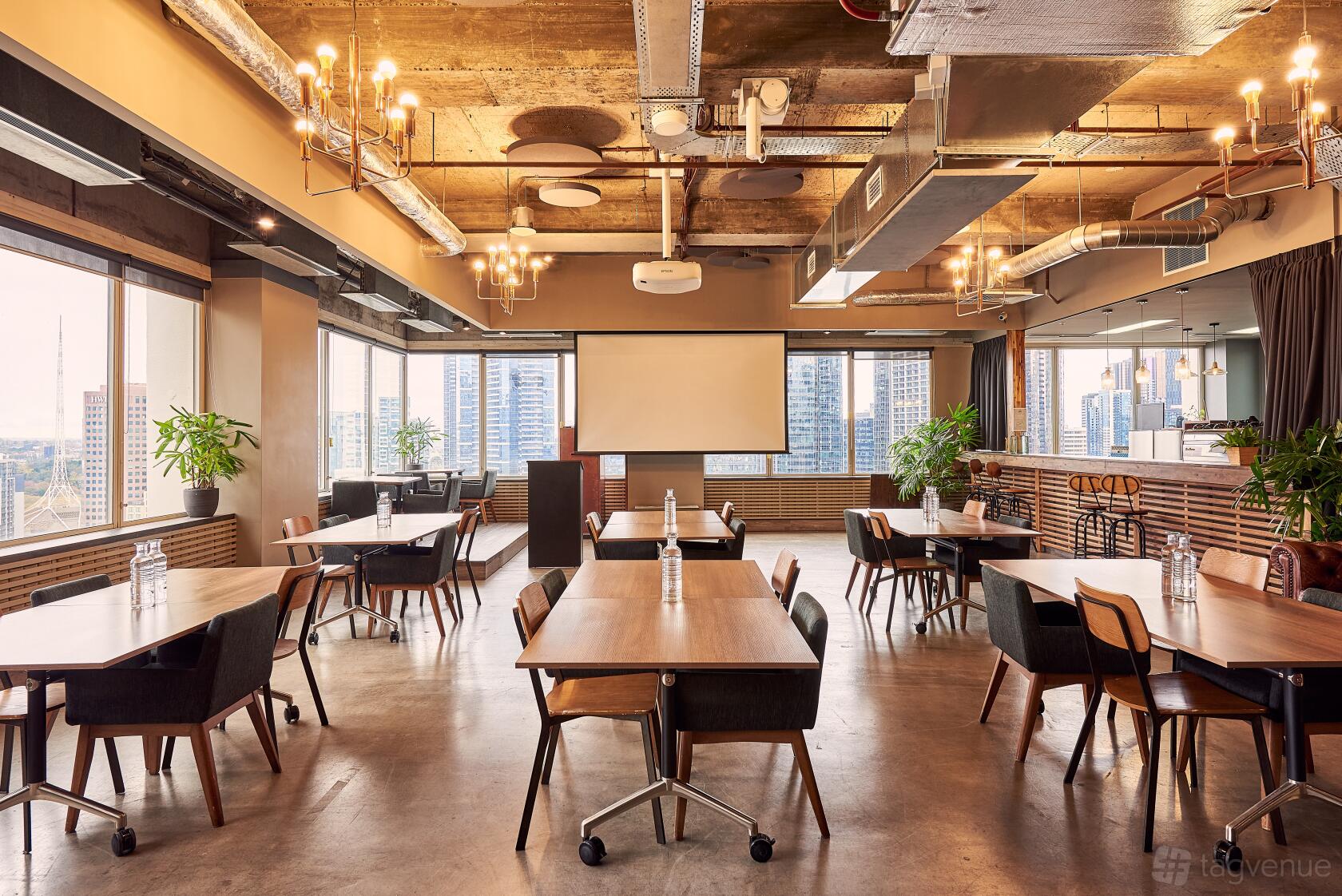 An event space with wooden tables, black chairs, chandeliers, and floor-to-ceiling windows overlooking the city at The Cluster.