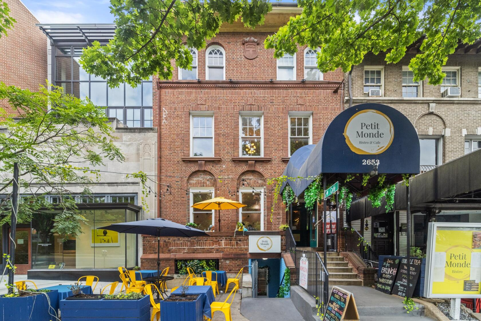 A restaurant with yellow patio chairs, blue planters, and large umbrellas outside Petit Monde Cafe.