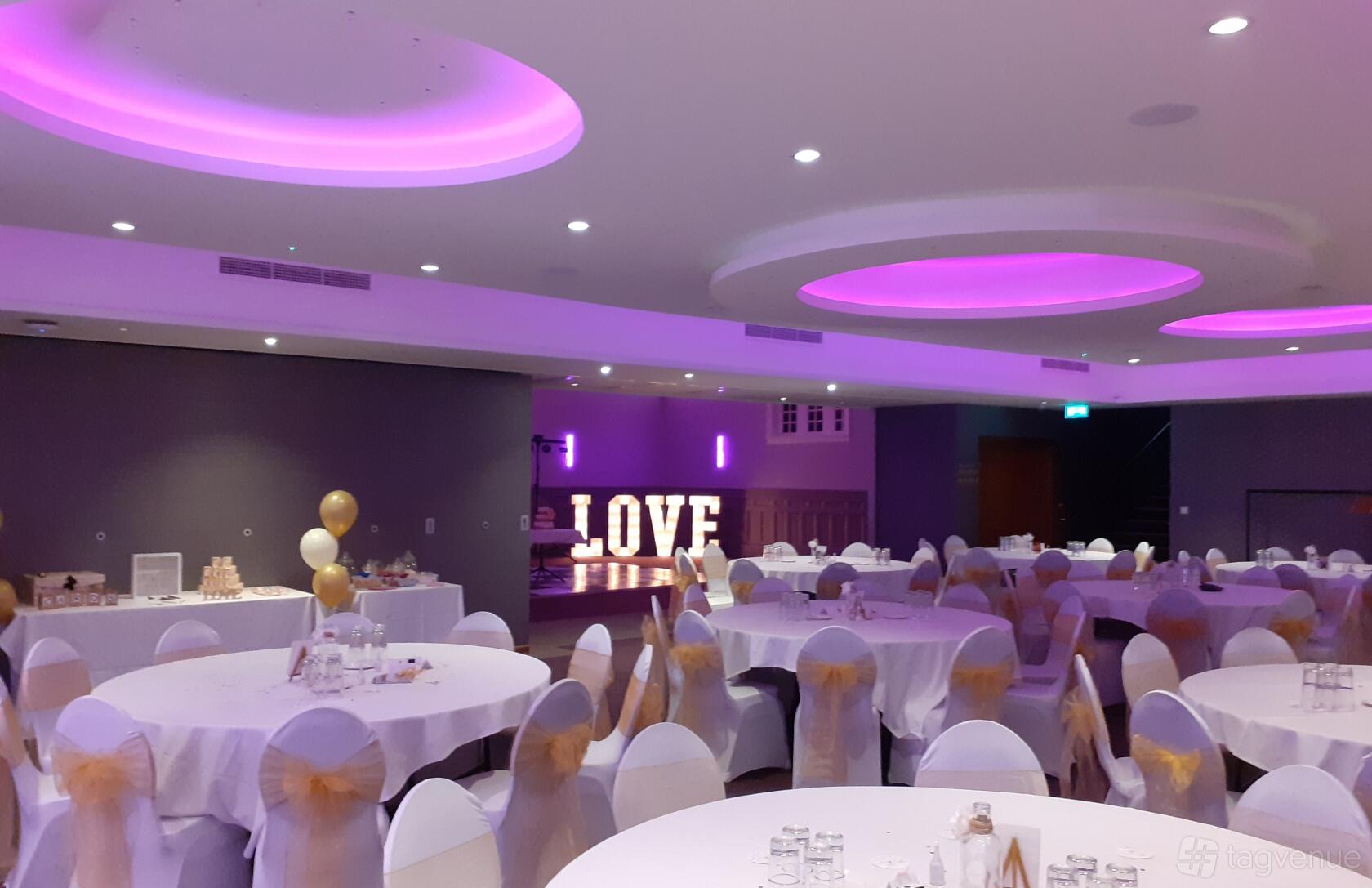An event space with round tables, white chair covers with gold bows, and purple ceiling lighting at The Place Hotel.